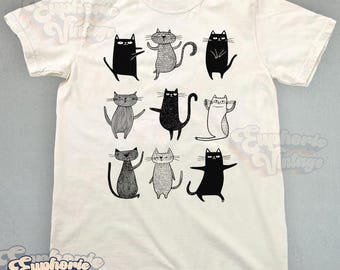 May include: A cream-colored short-sleeve t-shirt featuring a graphic print of nine stylized cats in black, white, and gray. The cats are depicted in various poses and textures, arranged in a three-by-three grid pattern on the front of the shirt.