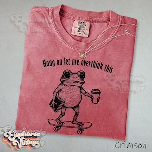Puede incluir: Una camiseta de manga corta color carmes&iacute; con un gr&aacute;fico negro de una rana en un monopat&iacute;n sosteniendo un libro y una taza de caf&eacute;. El texto sobre la rana dice "Hang on let me overthink this". La camiseta tiene un cuello redondo cl&aacute;sico.