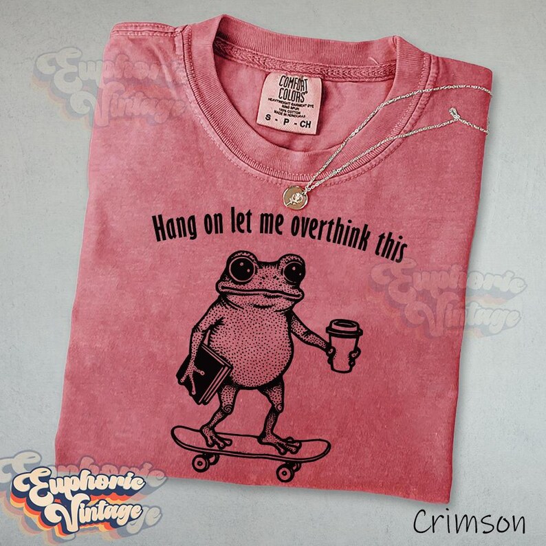 Puede incluir: Una camiseta de manga corta color carmes&iacute; con un gr&aacute;fico negro de una rana en un monopat&iacute;n sosteniendo un libro y una taza de caf&eacute;. El texto sobre la rana dice "Hang on let me overthink this". La camiseta tiene un cuello redondo cl&aacute;sico.