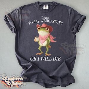 May include: Dark gray t-shirt featuring a cartoon frog wearing a pink cowboy hat and scarf, holding a coffee cup. Text reads "I Have To Say Weird Stuff Or I Will Die."