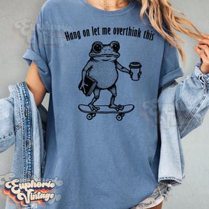 Puede incluir: Una camiseta azul con un estampado gr&aacute;fico negro de una rana en un monopat&iacute;n sosteniendo un libro y una taza de caf&eacute;. Sobre la rana, el texto dice, Hang on let me overthink this. La camiseta tiene mangas cortas y un cuello redondo.