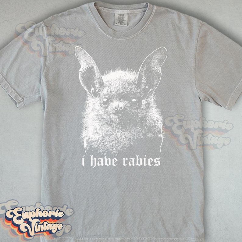 May include: A light gray t-shirt featuring a white illustration of a bat's face with large ears. Below the bat, the text reads "i have rabies". The shirt has a crew neck and short sleeves.