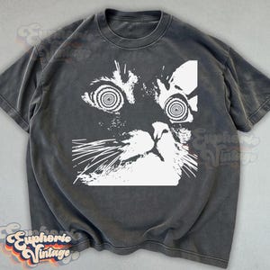 May include: Dark gray t-shirt featuring a white graphic of a cat's face with hypnotic spiral eyes. The cat's face is stylized with a geometric design. The shirt has a vintage, worn look.