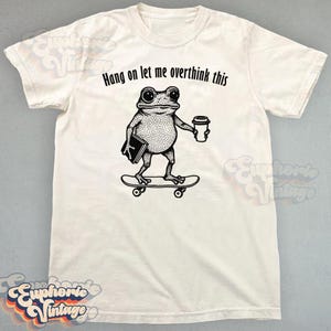 Puede incluir: Una camiseta de manga corta color hueso con un gr&aacute;fico negro de una rana sobre un monopat&iacute;n sosteniendo un libro y una taza de caf&eacute;. Sobre la rana, el texto "Hang on let me overthink this" aparece en una fuente negra y redondeada.