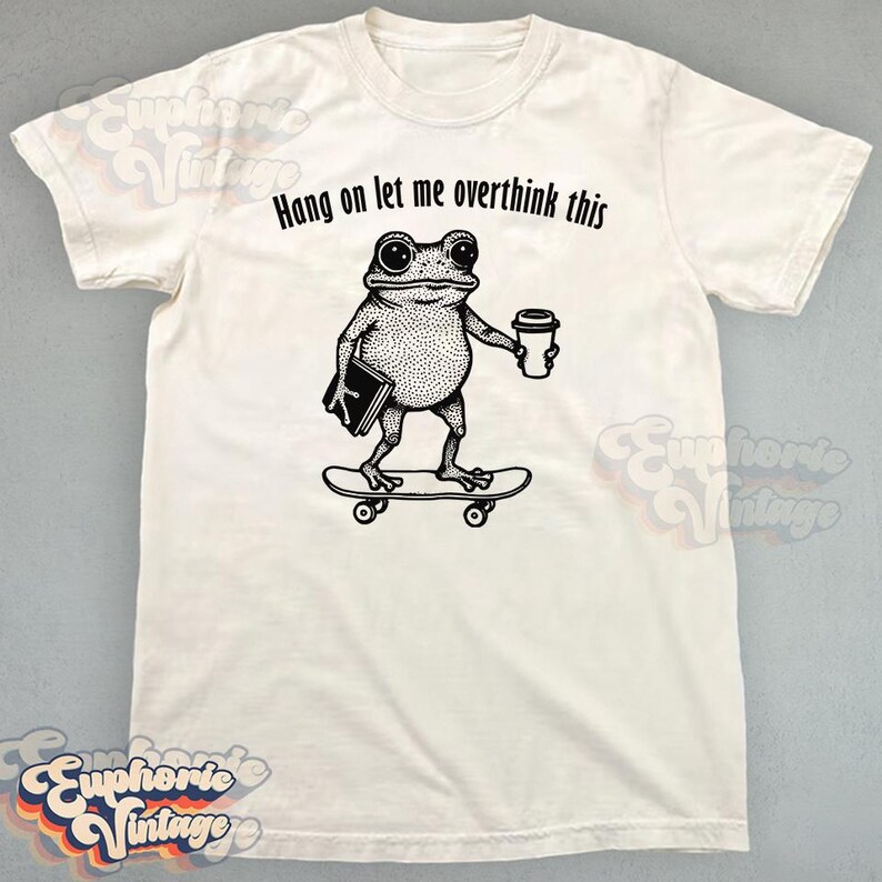 Puede incluir: Una camiseta de manga corta color hueso con un gr&aacute;fico negro de una rana sobre un monopat&iacute;n sosteniendo un libro y una taza de caf&eacute;. Sobre la rana, el texto "Hang on let me overthink this" aparece en una fuente negra y redondeada.