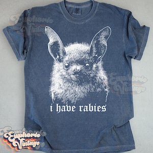May include: A blue t-shirt featuring a detailed white illustration of a bat's face. Below the bat, the text reads "i have rabies" in a gothic font. The shirt has short sleeves and a crew neck.