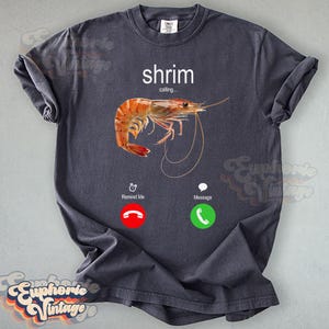 Op de afbeelding: Donkergrijs T-shirt met een aquarel illustratie van een garnaal en de tekst "shrim calling..." erboven. Onder de garnaal staan twee telefooniconen met de woorden "Remind Me" en "Message".