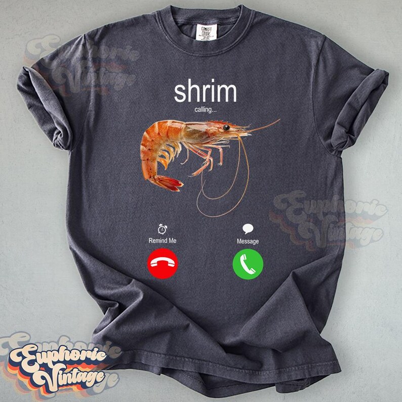 Op de afbeelding: Donkergrijs T-shirt met een aquarel illustratie van een garnaal en de tekst "shrim calling..." erboven. Onder de garnaal staan twee telefooniconen met de woorden "Remind Me" en "Message".