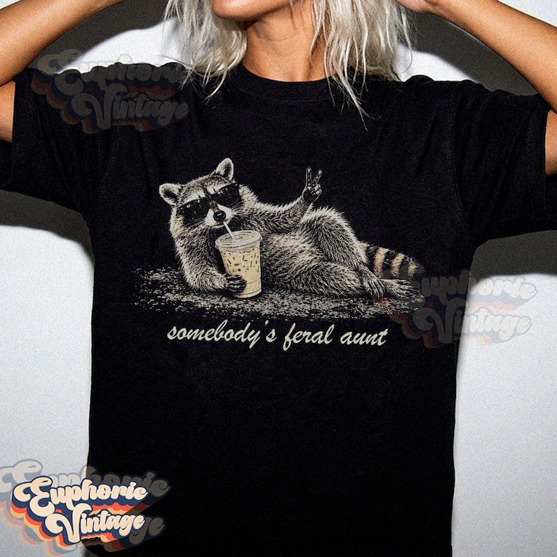 May include: A black t-shirt featuring a graphic of a raccoon wearing sunglasses, lying down while holding an iced coffee and making a peace sign. Below the raccoon, the text reads, somebody's feral aunt, in a white, lowercase, cursive font.