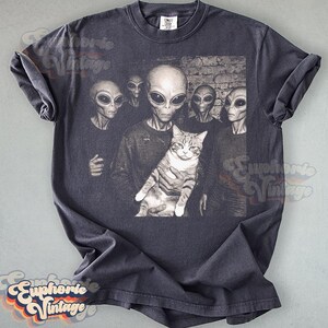 May include: Dark gray t-shirt featuring a group of aliens holding a cat. The aliens have large black eyes and gray skin. The cat is gray and white. The shirt has a vintage look.
