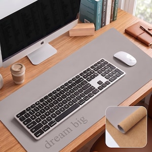 May include: A rectangular light grey desk mat featuring the text "dream big" in a serif font. The mat has a smooth top surface and a tan cork underside, shown in an inset image. A silver and black computer keyboard and a white mouse sit on the mat.