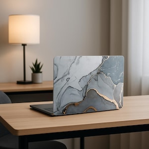 Może przedstawiać: Laptop z dekoracyjną obudową z szarym, białym i złotym abstrakcyjnym wzorem. Laptop jest otwarty na jasnym drewnianym biurku. Lampa i mała roślina znajdują się w tle.