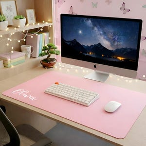 May include: A large, rectangular, light pink desk mat featuring the name Olivia printed in white script on the left side. The mat is positioned on a wooden desk surface, serving as a base for a white mechanical keyboard and a white computer mouse.