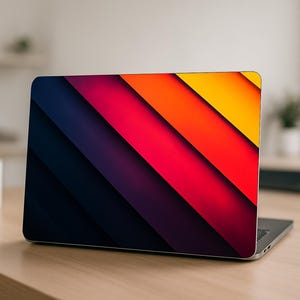 May include: Laptop with a vibrant diagonal design. The cover displays a gradient of colours, including dark blue, purple, red, orange, and yellow. The laptop is open on a wooden desk, with a white mug and a computer monitor in the background.