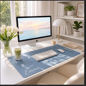 May include: A rectangular blue desk mat featuring the name Emma printed in white script lettering. The mat is positioned on a white desk surface, serving as a base for a white computer keyboard and a white computer mouse, creating a workspace setup.