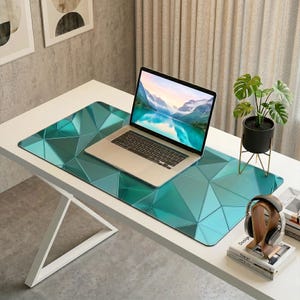 Puede incluir: Un ordenador portátil descansa sobre una alfombrilla de escritorio con estampado geométrico verde azulado. La alfombrilla tiene un patrón triangular repetido. Una pequeña planta en maceta y unos auriculares también están en el escritorio.