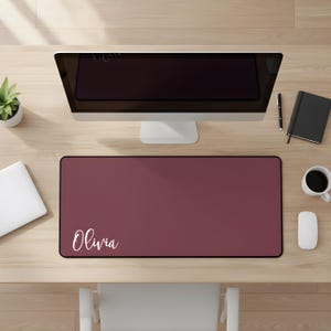 May include: A burgundy desk mat with the name "Olivia" in white script. The mat is rectangular and sits on a light wood desk, with a computer monitor, laptop, and coffee cup visible.