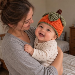 May include: A crochet baby hat shaped like an orange pumpkin with a green stem and green leaves on top. The hat features a textured ribbed design in bright orange yarn with a dark brown stem and two textured green leaves attached at the crown.