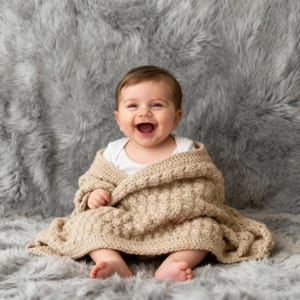 Könnte beinhalten: Ein lächelndes Baby, eingehüllt in eine beige Strickdecke. Das Baby trägt einen weißen Body und sitzt auf einer grauen, flauschigen Oberfläche. Die Decke hat ein strukturiertes Muster.