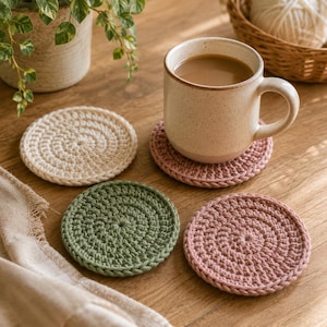 May include: A set of three round, crocheted coasters in cream, sage green, and dusty rose colors. One dusty rose coaster supports a speckled ceramic mug filled with coffee. The coasters feature a dense, circular stitch pattern with a braided edge.