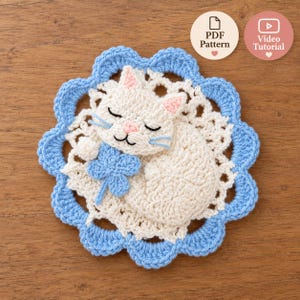 May include: A round crochet coaster featuring a cream-colored sleeping cat with pink ears and nose, holding a light blue four-leaf clover. The cat rests on a white lacy doily base with a scalloped light blue border. Text reads PDF Pattern and Video Tutorial.