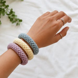 May include: Three circular bracelets featuring a textured, woven knit pattern in soft pastel colors. The set includes one light blue, one pale yellow, and one light purple bracelet, each with a thick, rounded profile and a consistent, uniform stitch.