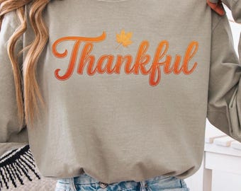 May include: A long-sleeved, Sandstone-colored shirt with the word "Thankful" in an orange and red gradient script. A small orange leaf graphic is above the word. The shirt is paired with denim shorts.