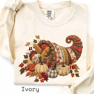 May include: Ivory-colored long-sleeve sweatshirt featuring a cornucopia design filled with pumpkins, gourds, corn, and fall leaves. The cornucopia is made of various patterned fabrics. The word "Ivory" is printed at the bottom of the shirt.