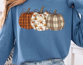 May include: A long-sleeved, blue jean colored sweatshirt featuring three decorative pumpkins. The pumpkins are in shades of orange, white, and brown, with different patterns including plaid and floral designs. The sweatshirt is a comfortable, casual style.