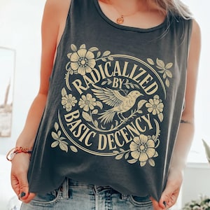 以下が含まれることがあります： 「RADICALIZED BY BASIC DECENCY」の文字と鳥、花の装飾が施された、ダークグレーのタンクトップ。タンクトップはノースリーブで、柔らかい素材でできています。