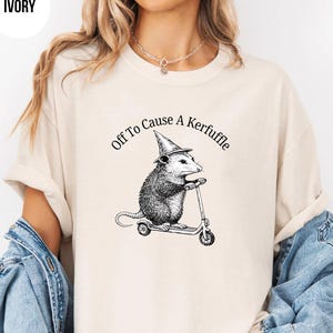 Könnte beinhalten: Elfenbeinfarbenes T-Shirt mit dem Text "Off To Cause A Kerfuffle". Die Grafik zeigt ein Opossum mit Hexenhut, das Roller fährt. Das Design ist in schwarzer Tinte.