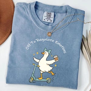 Puede incluir: Camiseta azul claro con la frase "Off To Regulate Emotions" que rodea a un pato de dibujos animados montado en un scooter y sosteniendo una taza de café. La camiseta tiene cuello redondo y mangas cortas. Un collar dorado está drapeado sobre la camiseta.