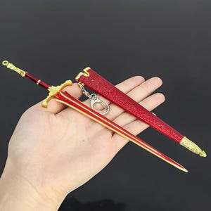May include: A miniature sword keychain with a red blade and scabbard, accented with gold-colored details. The sword features a cross-shaped hilt and a decorative handle. The scabbard is textured and includes a keyring attachment.