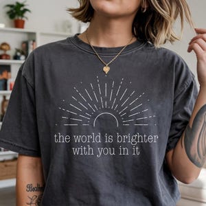 Peut inclure: T-shirt gris foncé avec un motif de soleil blanc et le texte "the world is brighter with you in it". Le t-shirt a un aspect vieilli. Une personne porte le t-shirt et un collier en or.