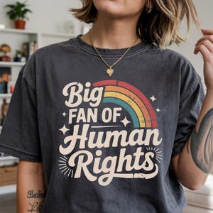 May include: Dark grey t-shirt with the slogan "Big Fan of Human Rights" in a retro font, featuring a rainbow design. The t-shirt has a vintage, washed appearance.