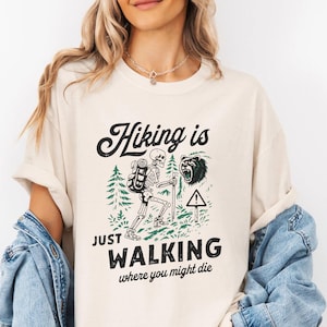 May include: Cream-colored t-shirt with the text "Hiking is just walking where you might die." The graphic features a skeleton hiker, a bear, and trees in green and black. The shirt has rolled-up sleeves.