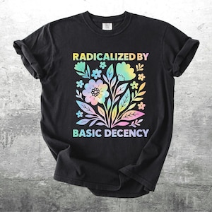 以下が含まれることがあります： 黒いTシャツ。カラフルな花の模様と「RADICALIZED BY BASIC DECENCY」の文字がプリントされています。パステルレインボーカラーの花と葉が描かれており、暗い生地とのコントラストが鮮やかです。