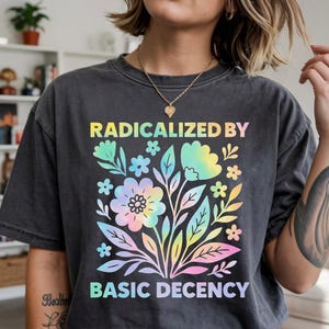 May include: Dark grey t-shirt with a colourful floral design and the text "RADICALIZED BY BASIC DECENCY". The design features flowers and leaves in pastel rainbow colours. The shirt has a vintage, washed look.