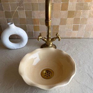 Peut inclure: Une vasque en céramique de couleur crème avec un bord festonné et ondulé et une bonde circulaire en laiton. La vasque est placée devant un grand robinet en laiton de style ancien avec deux poignées en croix et un bec courbé, finition lisse.