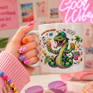 Peut inclure: Mug en céramique blanche avec un motif de serpent de dessin animé portant un chapeau et une chemise hawaïenne, tenant une glace et un taco. Le mug porte l'inscription "Summer Chaos Mashup Fri!" et "Pou: Chaos".