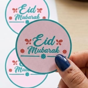 Peut inclure: Autocollants circulaires roses et turquoise avec l'inscription "Eid Mubarak" en script turquoise. Les autocollants ont une bordure turquoise et des motifs floraux. Une main aux ongles bleus tient un autocollant.