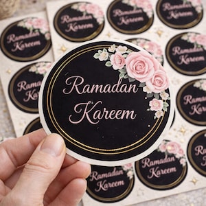 Peut inclure: Ronds autocollants noirs avec bordure dorée, portant l'inscription "Ramadan Kareem" en rose. Les autocollants sont ornés de roses roses et de petites fleurs blanches. Une main tient un autocollant.