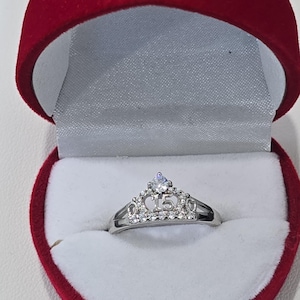 May include: A silver-toned ring shaped like a tiara, featuring a central number 15 and embedded with clear, sparkling stones. The ring is displayed inside a heart-shaped, red velvet jewelry box with a white interior lining and a hinged lid.