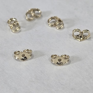 May include: Five gold-toned metal earring backs arranged in a circular pattern. Each earring back features a heart-shaped design with a central hole for an earring post and two curved metal loops that provide tension to secure the earring in place.