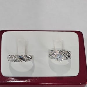 May include: A silver-toned ring set featuring a band with diagonal grooves and embedded stones, alongside a matching engagement ring with a central round stone and a coordinating band, both displayed on a white stand with a maroon base.