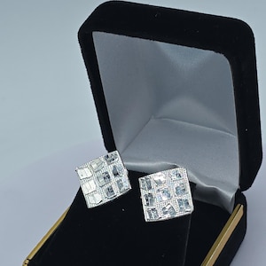 May include: A pair of square silver stud earrings featuring a grid pattern of small, reflective, multi-faceted metallic squares. The earrings are displayed inside a black velvet jewellery box with a gold-toned trim along the bottom edge.
