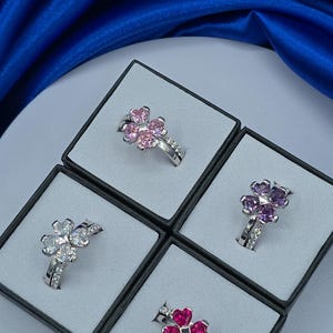 May include: Four silver-toned rings, each featuring a four-leaf clover design composed of four heart-shaped gemstones. The rings include one with light pink stones, one with clear stones, one with purple stones, and one with deep red stones.