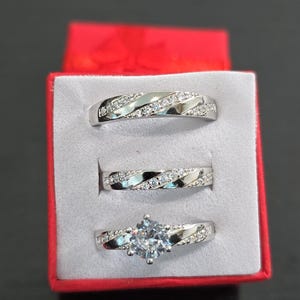 May include: A set of three silver-toned rings displayed in a red box. Two rings feature a diagonal channel of small clear stones, while the third ring includes a larger central clear stone flanked by two diagonal channels of small clear stones.