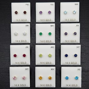 May include: Twelve pairs of 14K gold stud earrings with round faceted birthstones, each mounted on a white card labelled with months from ENE to DIC. The stones display various colours including red, purple, blue, green, yellow, and pink.