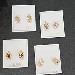 Puede incluir: Pendientes de tuerca dorados con forma de tortuga y diseños de concha de varios colores, incluyendo rosa, morado y rojo. Cada par se muestra en una tarjeta blanca con "14 K GOLD" impreso. Los pendientes están sobre una superficie gris oscuro.